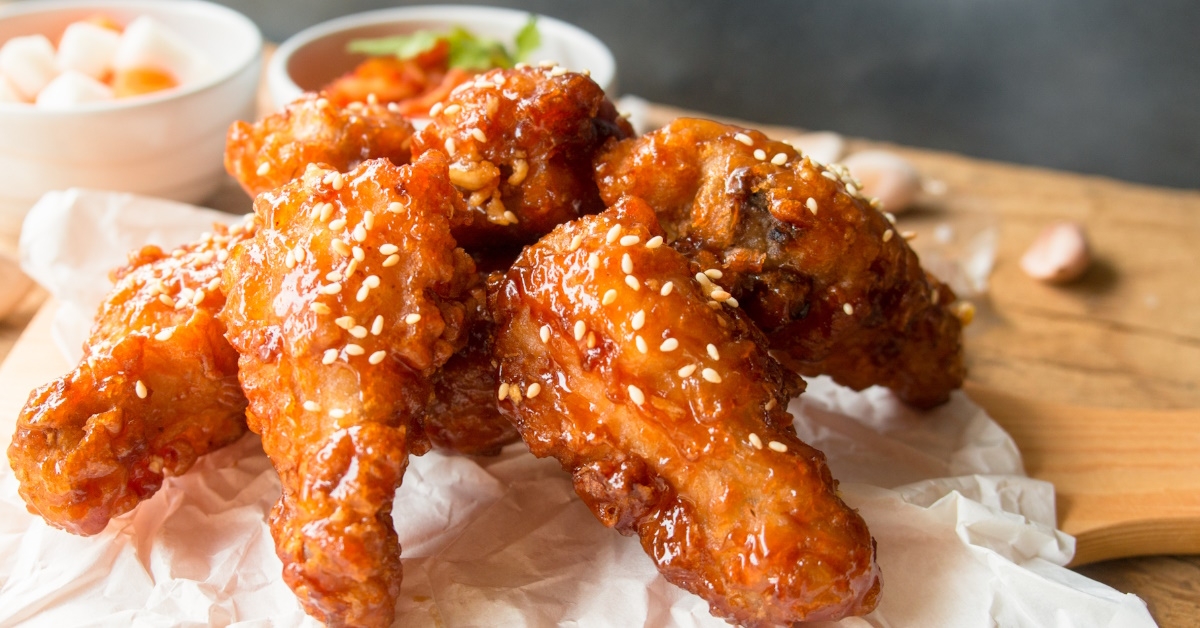 A wooden table displays a Korean-style deep-fried chicken wing with garlic sauce, kimchi, and pickled radish as accompaniments.