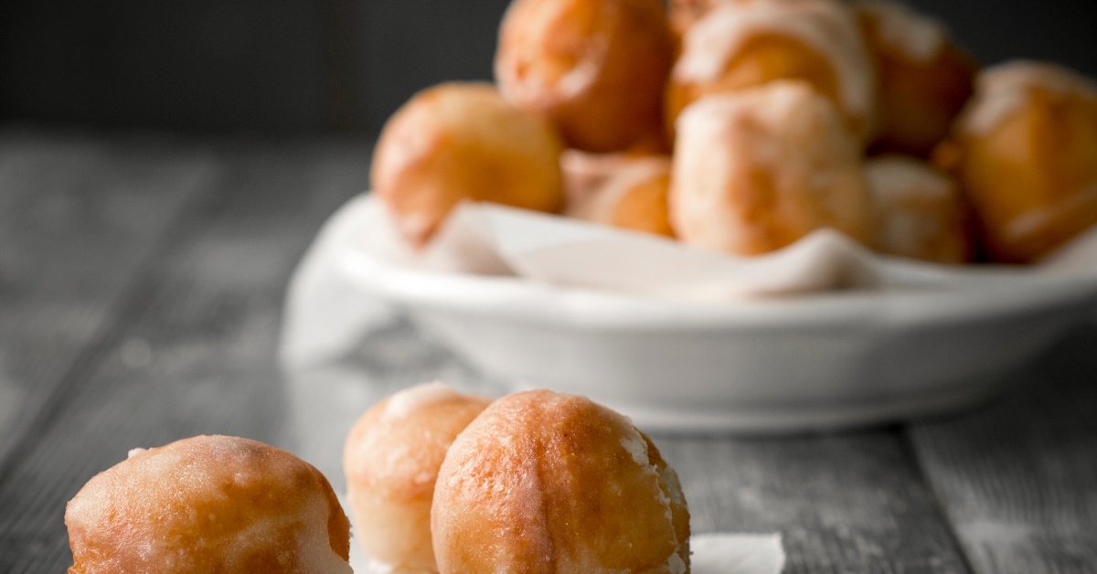 Three doughnuts holes are placed on a piece of baking paper in the foreground, with a white bowl filled with more doughnuts visible in the background.