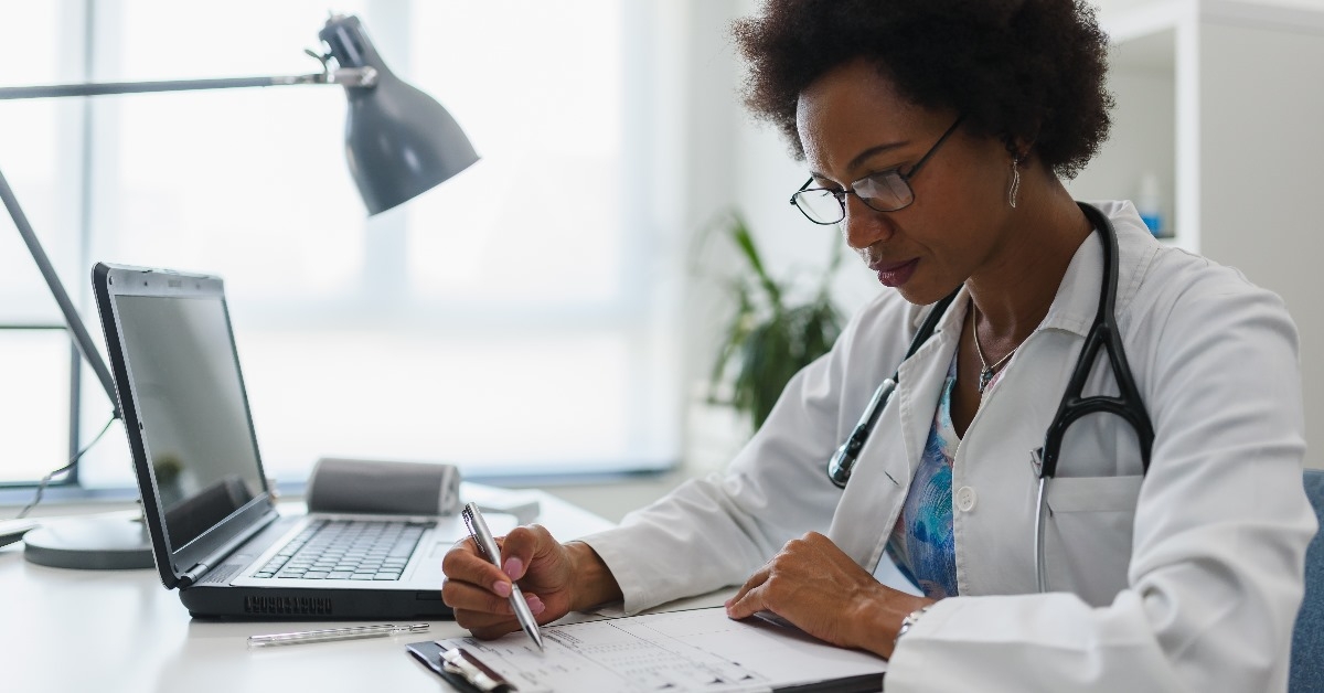 An African American doctor with short curly hairs carefully reviewing clinical reports of a patient while seated in her clinic with a focused expression.