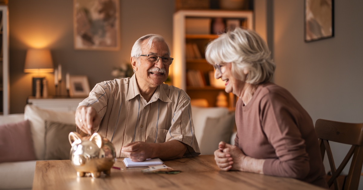 Elderly Couple Smiles At Table With Piggy Bank And Papers