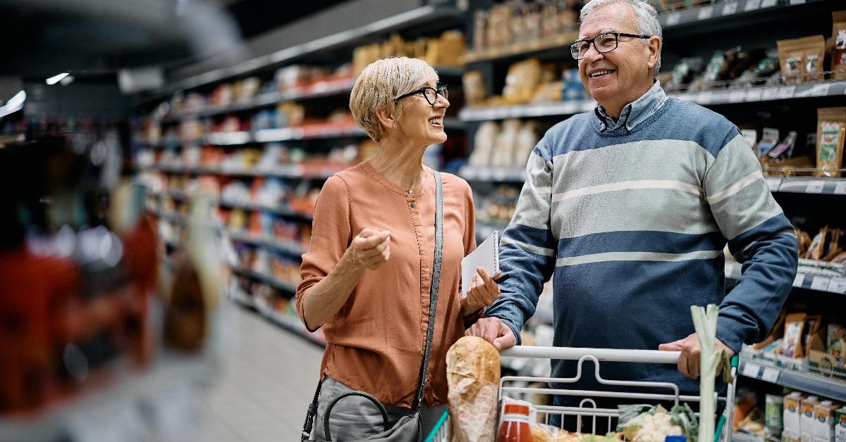 happy senior couple talking to each other while doing grocery with shopping trolley and notebook