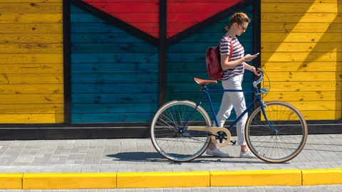 Woman walking her bike along sidewalk, looking down at phone