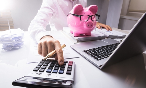 person calculating in front of a piggy bank and a laptop