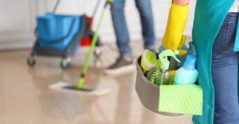 Female janitor holding a basket of cleaning supplies in the foreground with another janitor mopping in the background.l