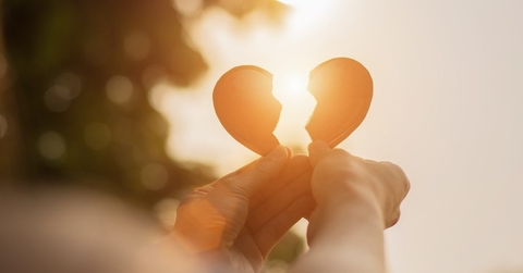 A pair of hands holding up two pieces of a broken heart toward the setting sun. 