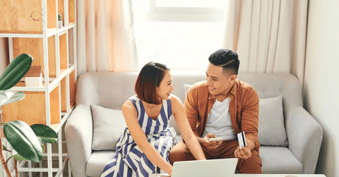 A couple sits on the couch together as they use the laptop in front of them and smile at one another.