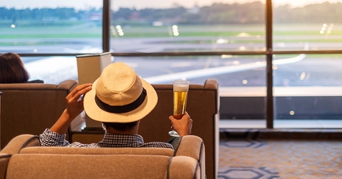 A man sits in a chair at the airport holding a beer and touching the brim of his hat. 
