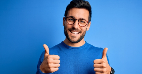young man in blue t shirt wearing glasses doing thumbs up in front of blue background