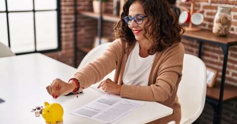 middle age woman sitting at table with papers putting pennie into yellow piggy bank