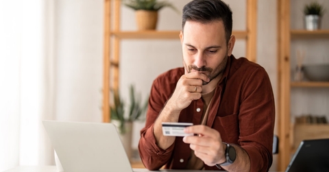 businessman sitting at table with laptop holding credit card thinking about loan at home