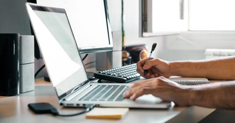a partially visible person shown working on a laptop while holding a pen in one hand