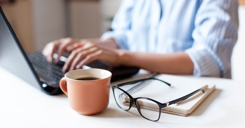 working on laptop with coffee, notebook, and glasses on table