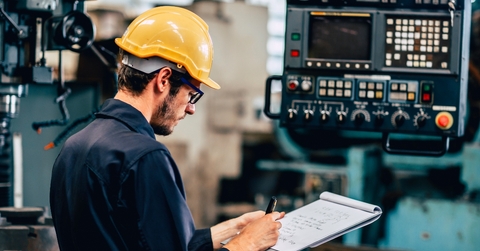 Man wearing yellow protection hat in a factory, taking notes 