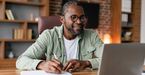 Man taking notes sitting at the desk looking at laptop