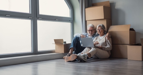 Older couple sitting against moving boxes with laptop